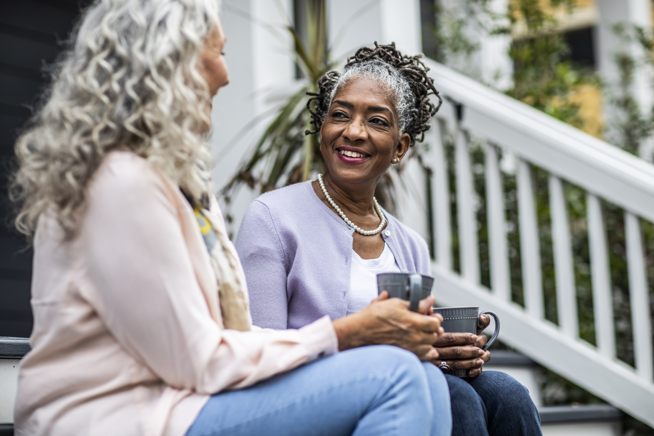 Two senior women hold coffee cups while chatting on the front porch