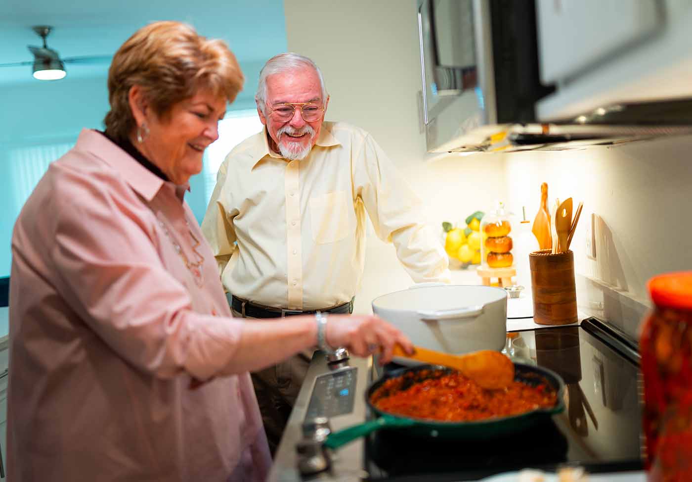 A senior couple stand in the kitchen while one of them stirs a pasta sauce in a pan over the stove-top range