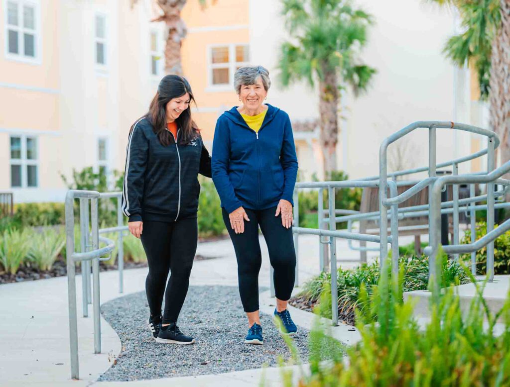 A senior woman and her physical therapist walk down a ramp in an outdoor courtyard