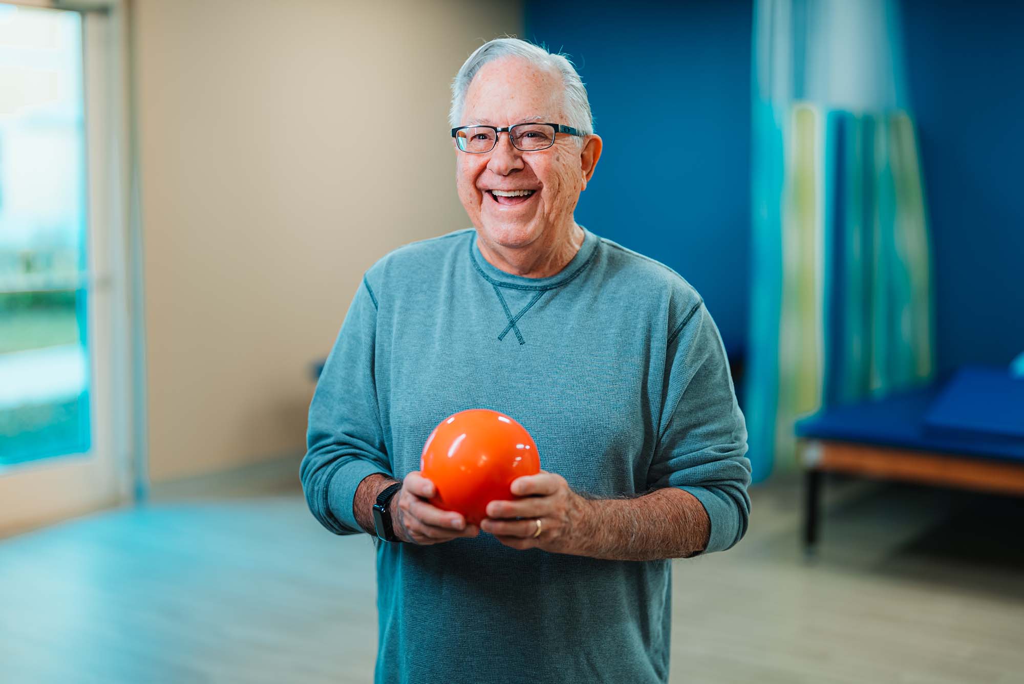 A senior man hold a physical therapy ball in an indoor rehabilitation room