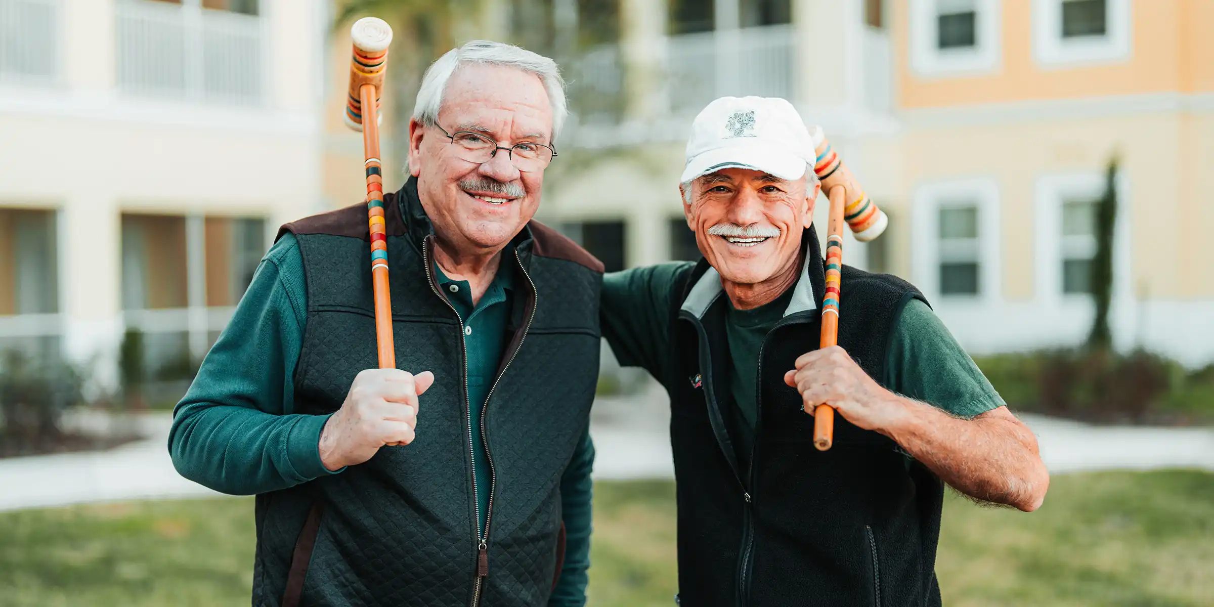 Two senior men pose with croquet mallets in an courtyard
