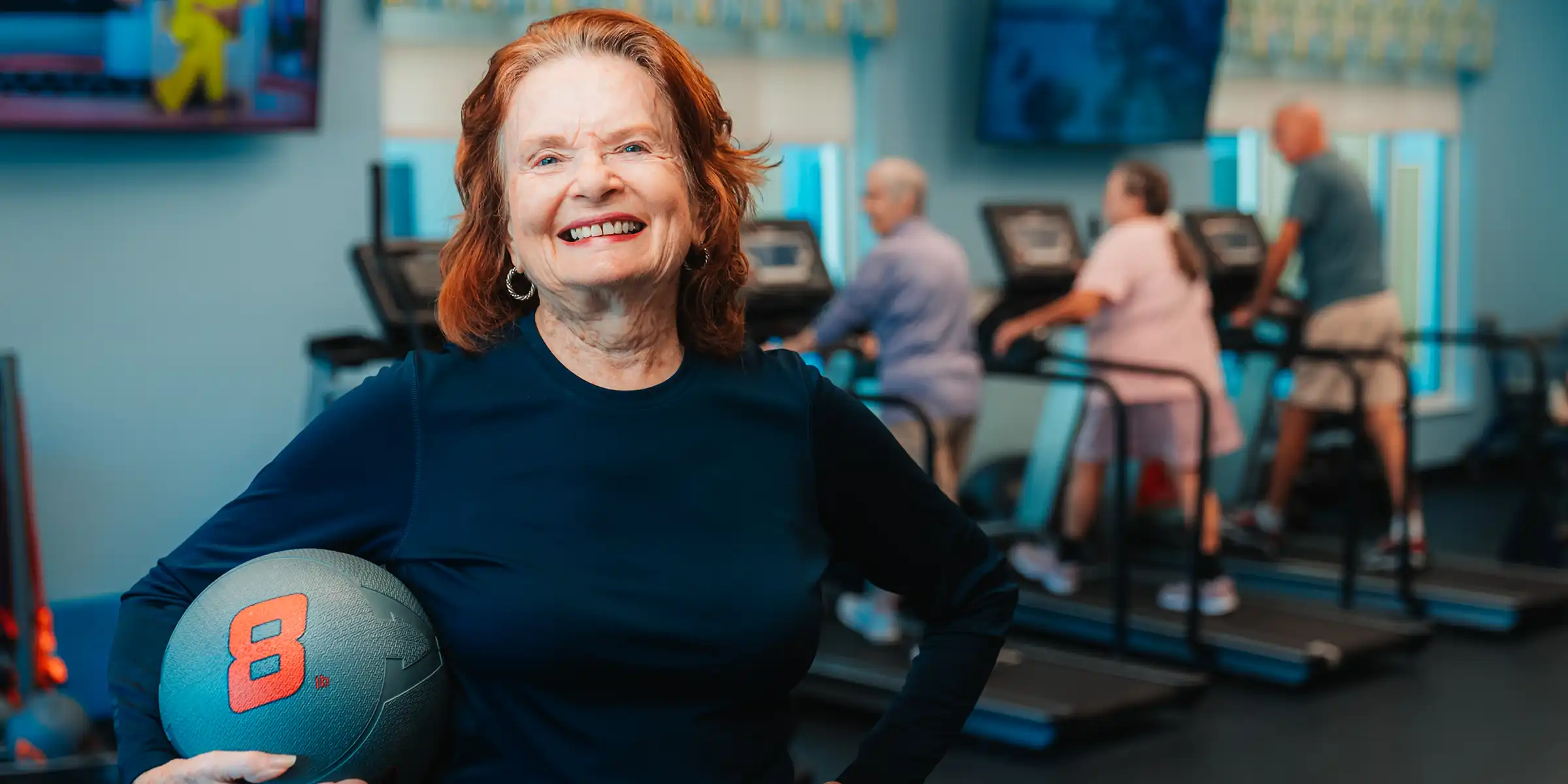 A senior woman poses with a weighted medicine ball in a wellness gym