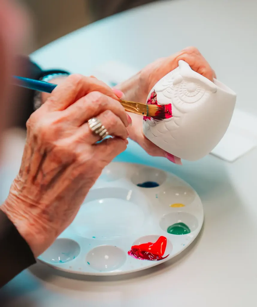 A close-up of a senior woman's hands paint on glaze of a ceramic owl pot