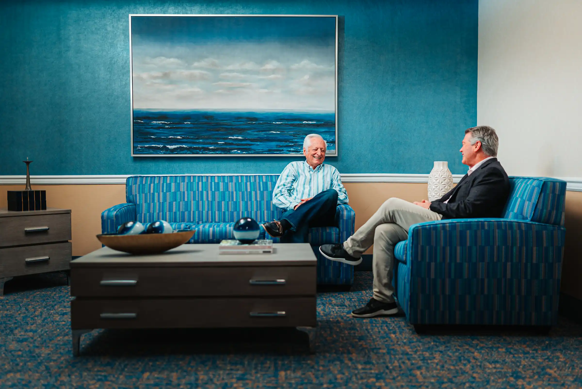 Two senior men sit and chat in a lounge area