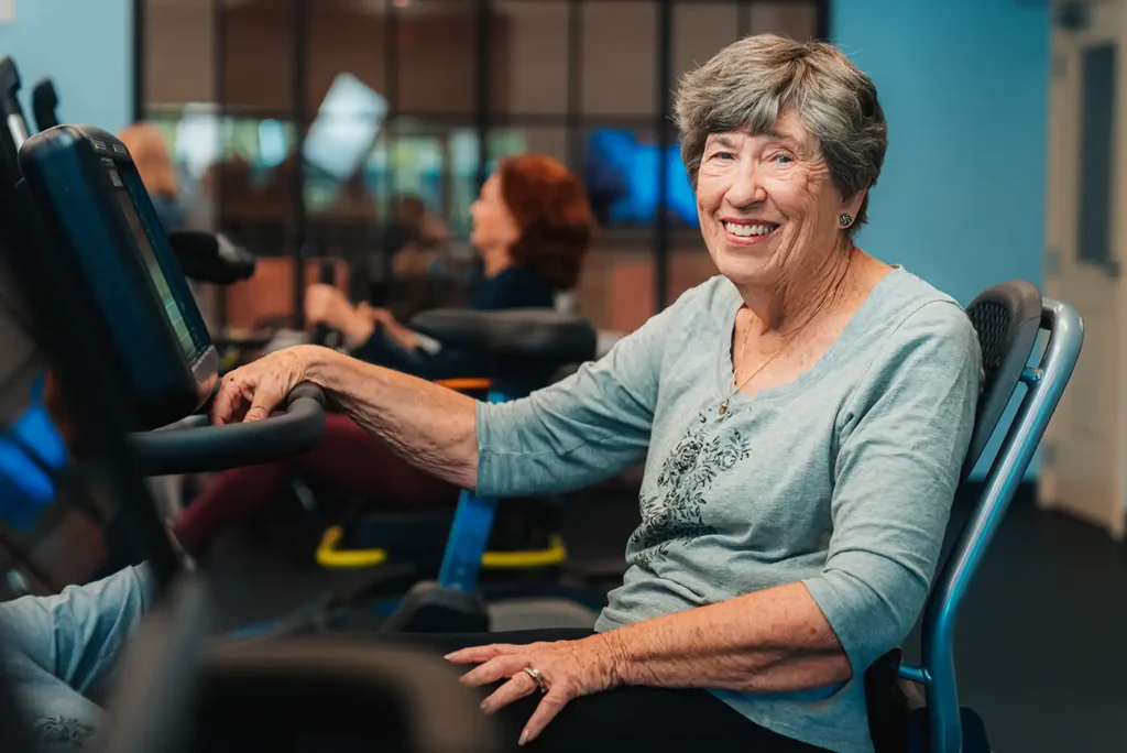 A senior woman poses seated on a piece of gym equipment in a workout room