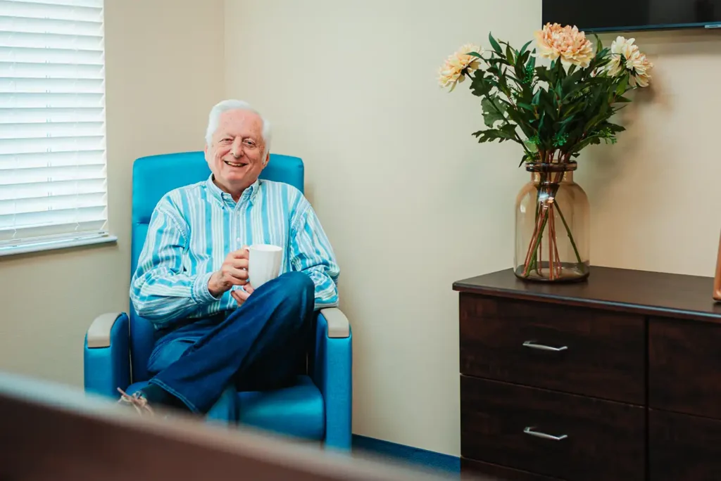 A senior man has coffee while sitting in a chair in the corner of a room